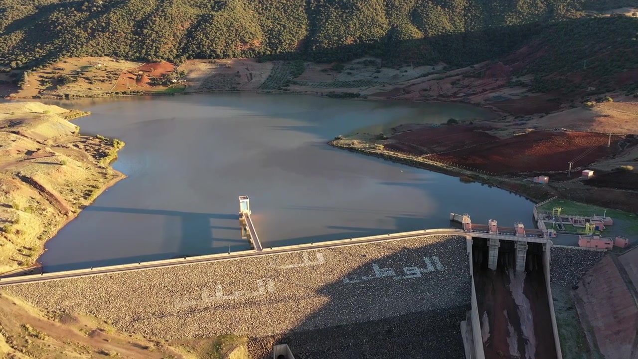 Vista aérea cinematográfica con dron de una presa de montaña y un embalse rodeados por un paisaje dramático en el Atlas Medio, Marruecos, ideal para proyectos de infraestructura y naturaleza.