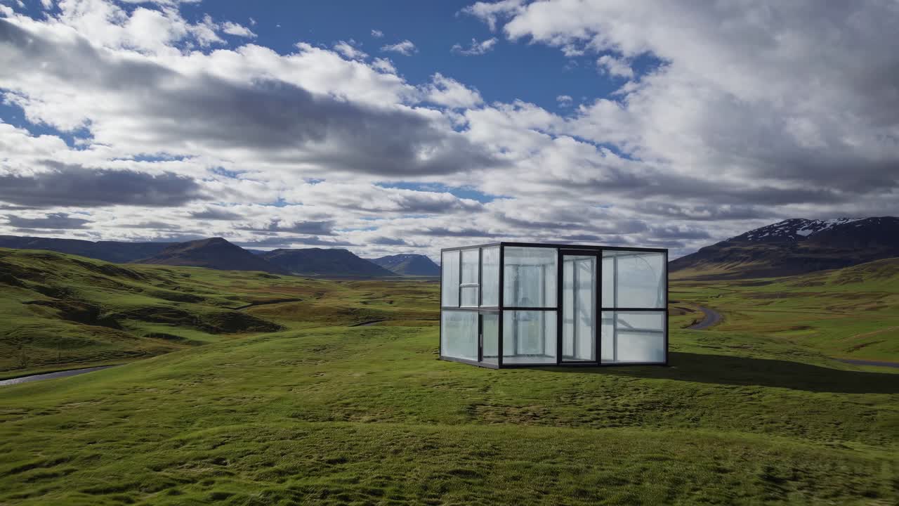 Glass house standing on a green hill in Iceland, illuminating the landscape under a cloudy sky, creating a contrast between nature and modern architecture