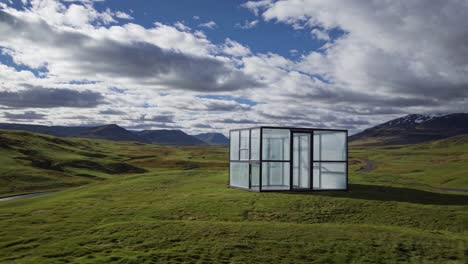 glass house standing on a green hill in iceland, illuminating the landscape under a cloudy sky, creating a contrast between nature and modern architecture