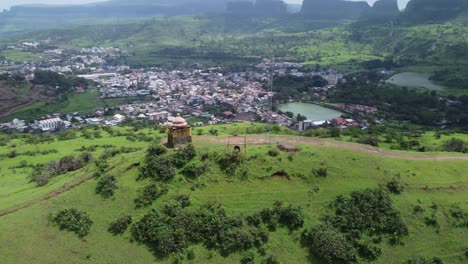 Drones-Dando-Vueltas-Alrededor-Del-Templo-Hindú-En-El-Borde-De-La-Cima-De-La-Montaña-Con-Vistas-A-La-Ciudad-De-Trimbkeshwar,-La-Presa-De-Ahilya-Y-Las-Cataratas-De-Trimbak-Al-Fondo-Rodeadas-De-Exuberante-Vegetación,-Nashik,-India