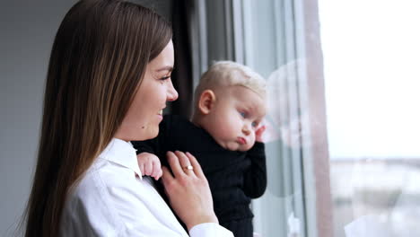 Loving Caucasian mother holds a baby standing at the window. Mom kisses her serious child looking outside.