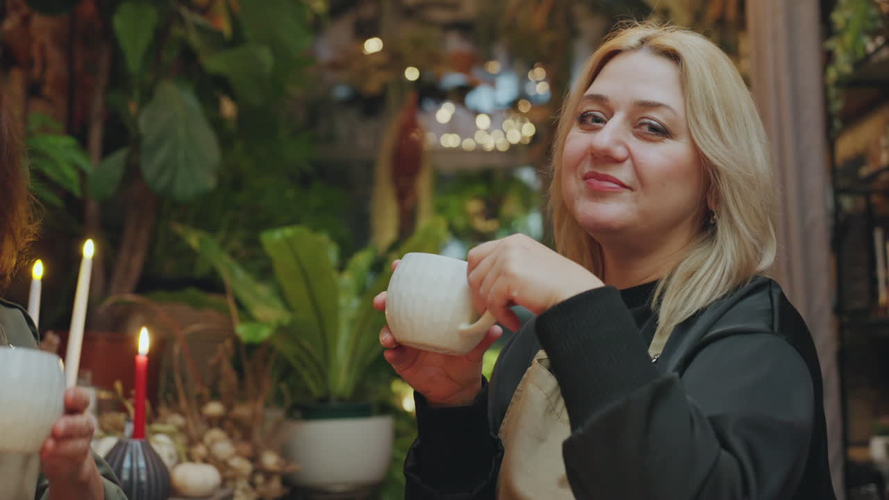 Florista caucásica rubia bebiendo de una taza de té, rodeada de exuberantes plantas y luz de velas, en una acogedora escena de tienda interior. Manos sosteniendo una taza de cerámica, sonrisa suave, ambiente relajado, estantes con jarrones, follaje y ramos.