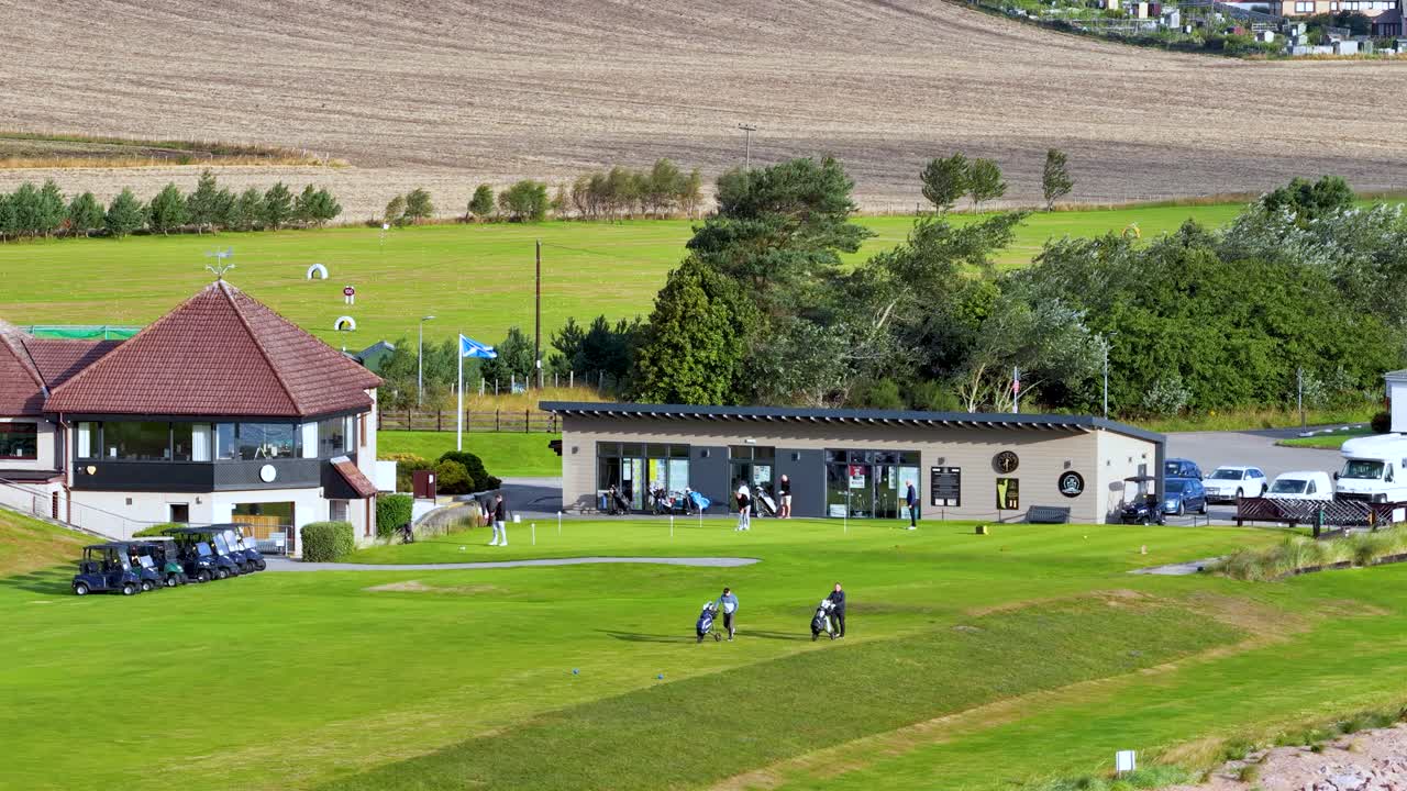 Two golfers walk on green fairway near clubhouse, sunny daylight, wide static shot, rural setting