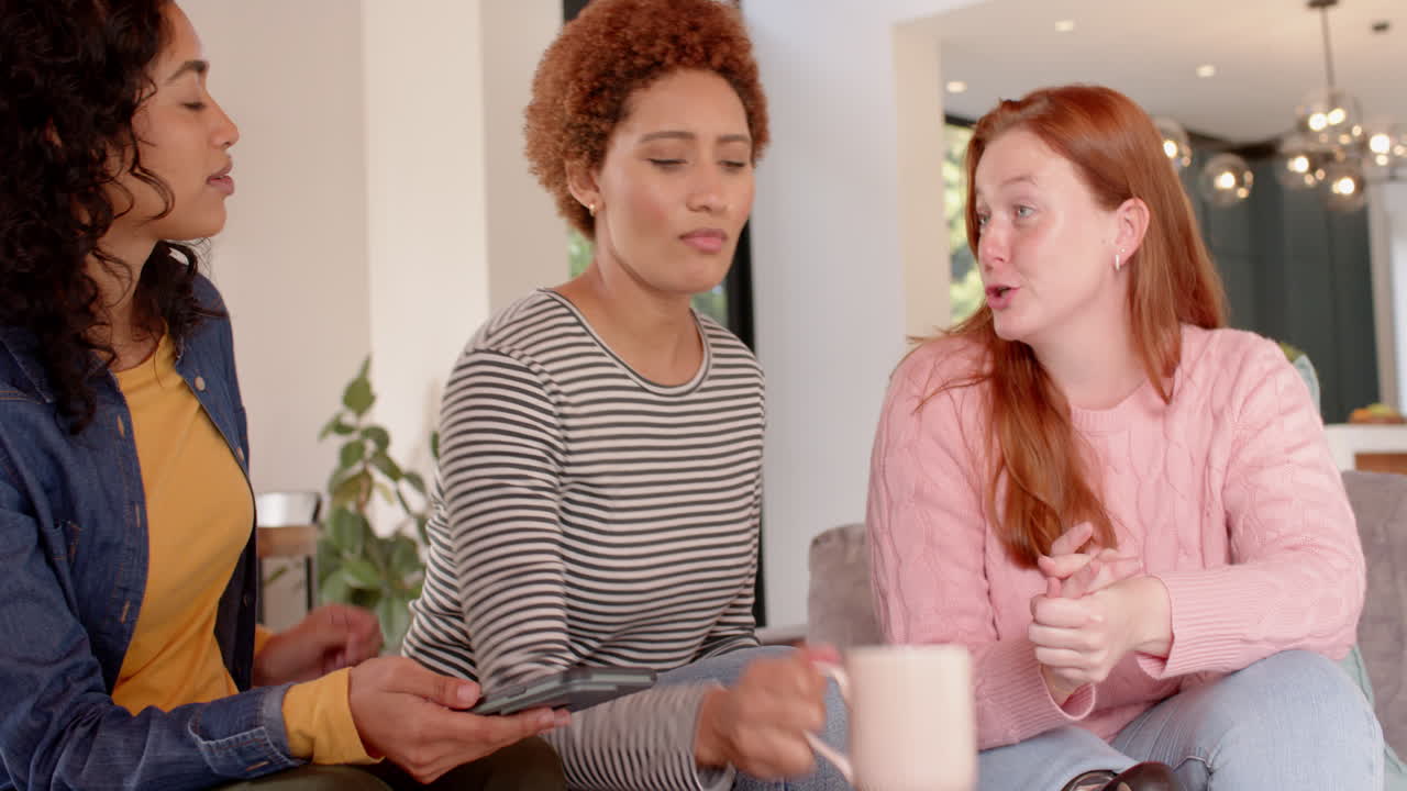 Chatting and laughing, diverse female friends hanging out together on couch indoors