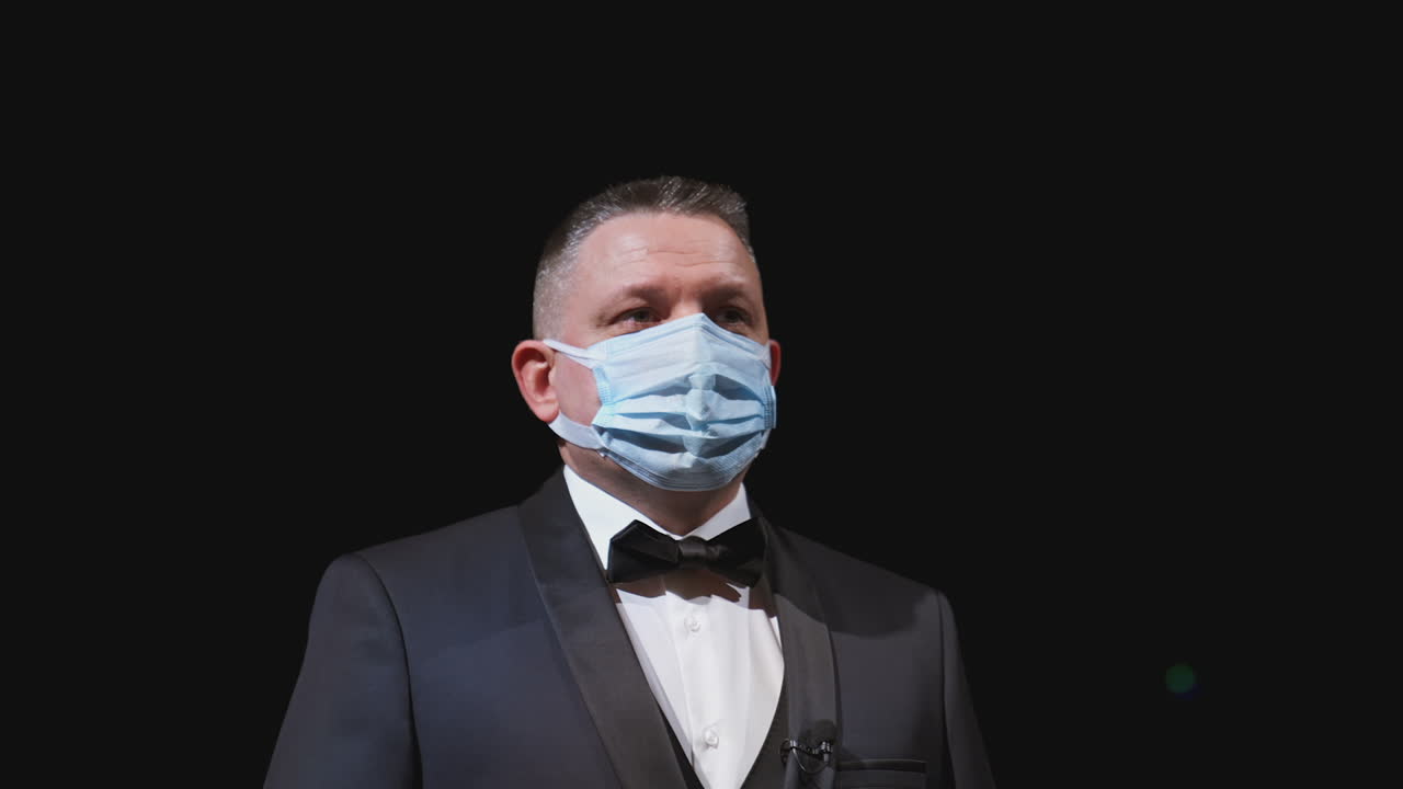 Man in elegant suit and medical mask. Portrait of an actor in protective facial mask standing on stage of a theater. Man putting off mask from face. Coronavirus pandemic.