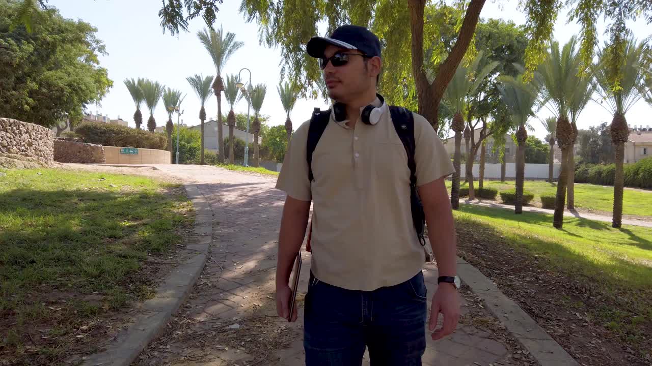 Front view of a young man with backpack and cap walking through a park.