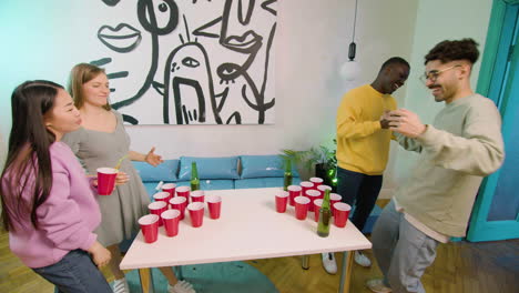 happy multiethnic young friends playing beer pong at home