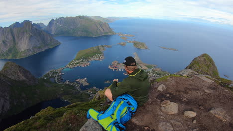 Hombre-Viajero-Sentado-En-Una-Colina-Del-Hermoso-Fiordo-Noruego-Reinebringen-Cerca-De-La-Ciudad-De-Reine,-Islas-Lofoten