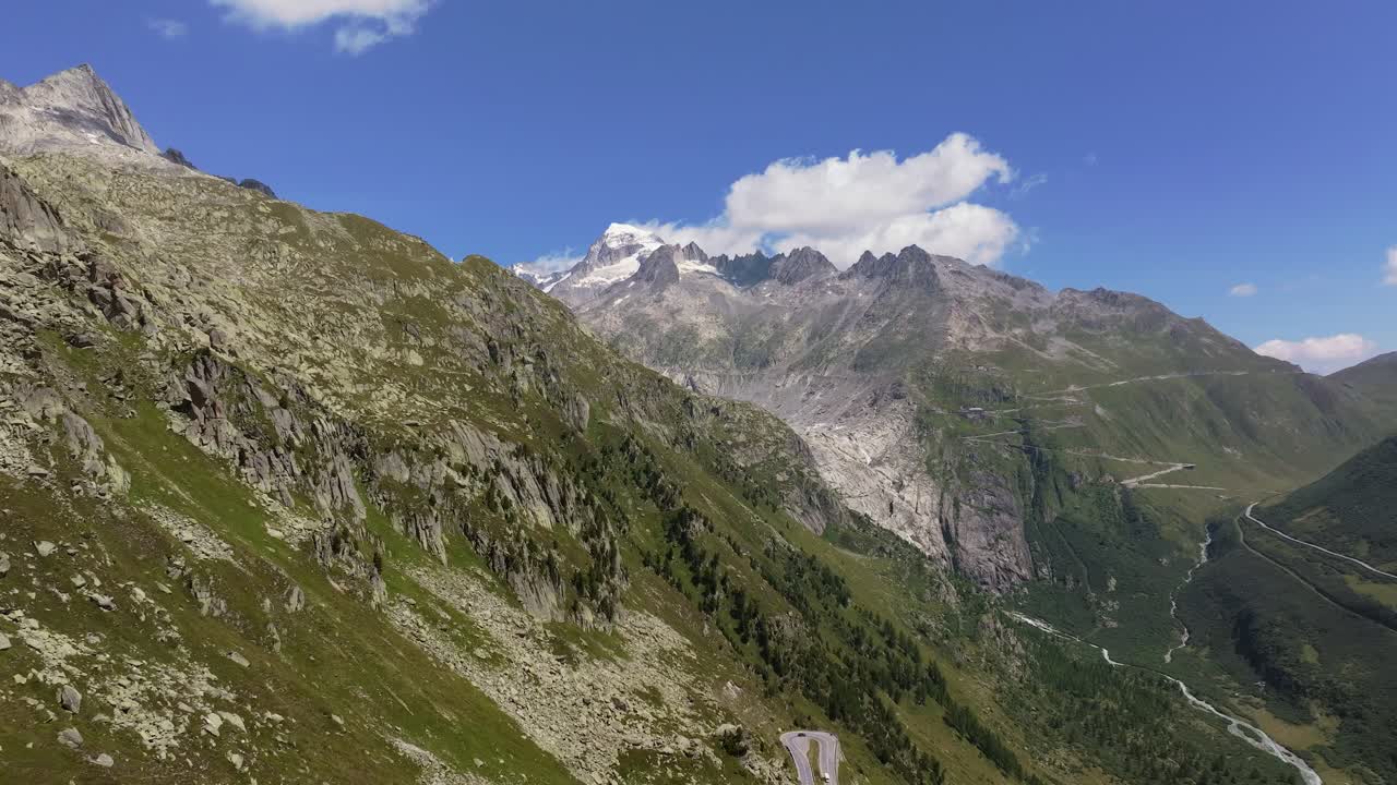 Aerial shot of a vast alpine valley surrounded by steep cliffs, green slopes, and distant snow-capped peaks, beneath a clear summer sky