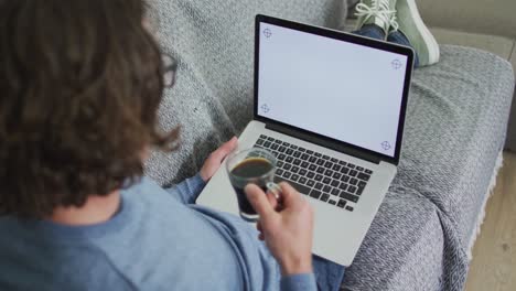 Caucasian-man-sitting-on-sofa-in-living-room-using-laptop-and-drinking-coffee