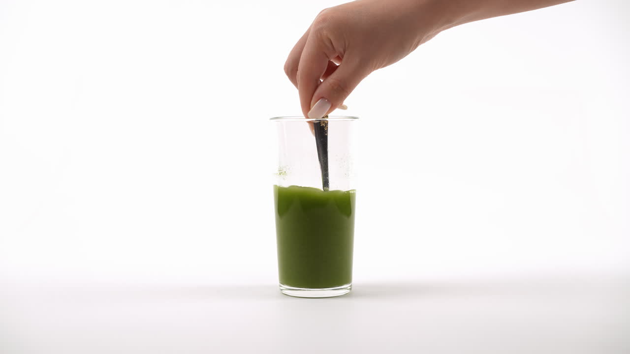 la mano agitando un batido verde con un tenedor en un vaso transparente contra un fondo blanco