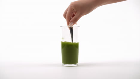 la mano agitando un batido verde con un tenedor en un vaso transparente contra un fondo blanco