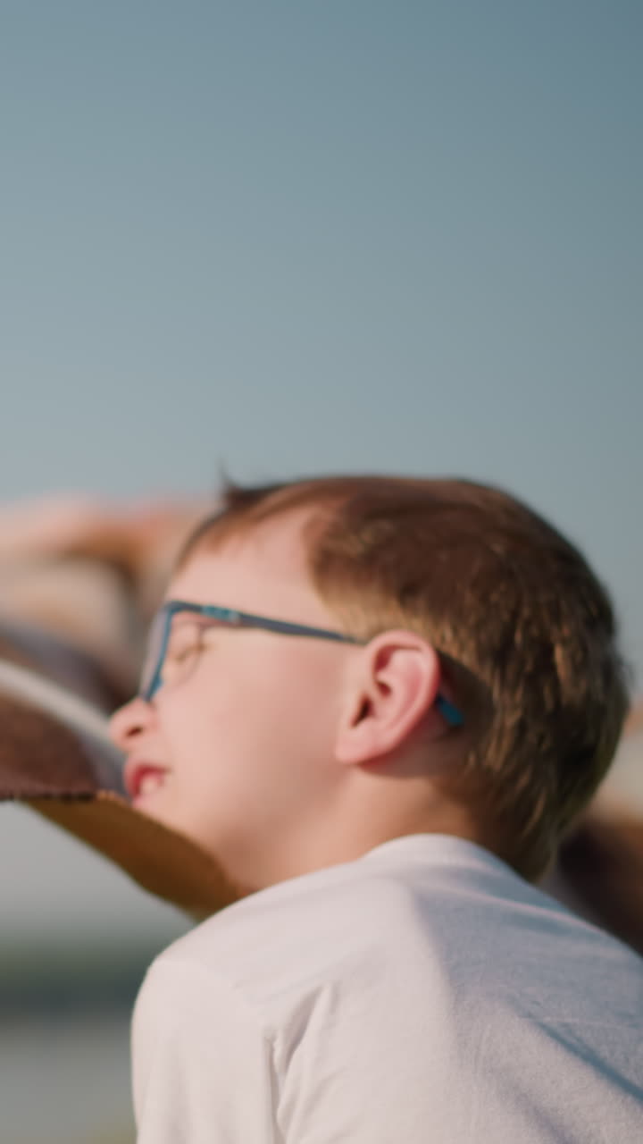 A young boy in a white shirt and glasses holds a plaid scarf in a grassy field beside a lake. In the background, a blurred woman in a blue dress helps with the scarf