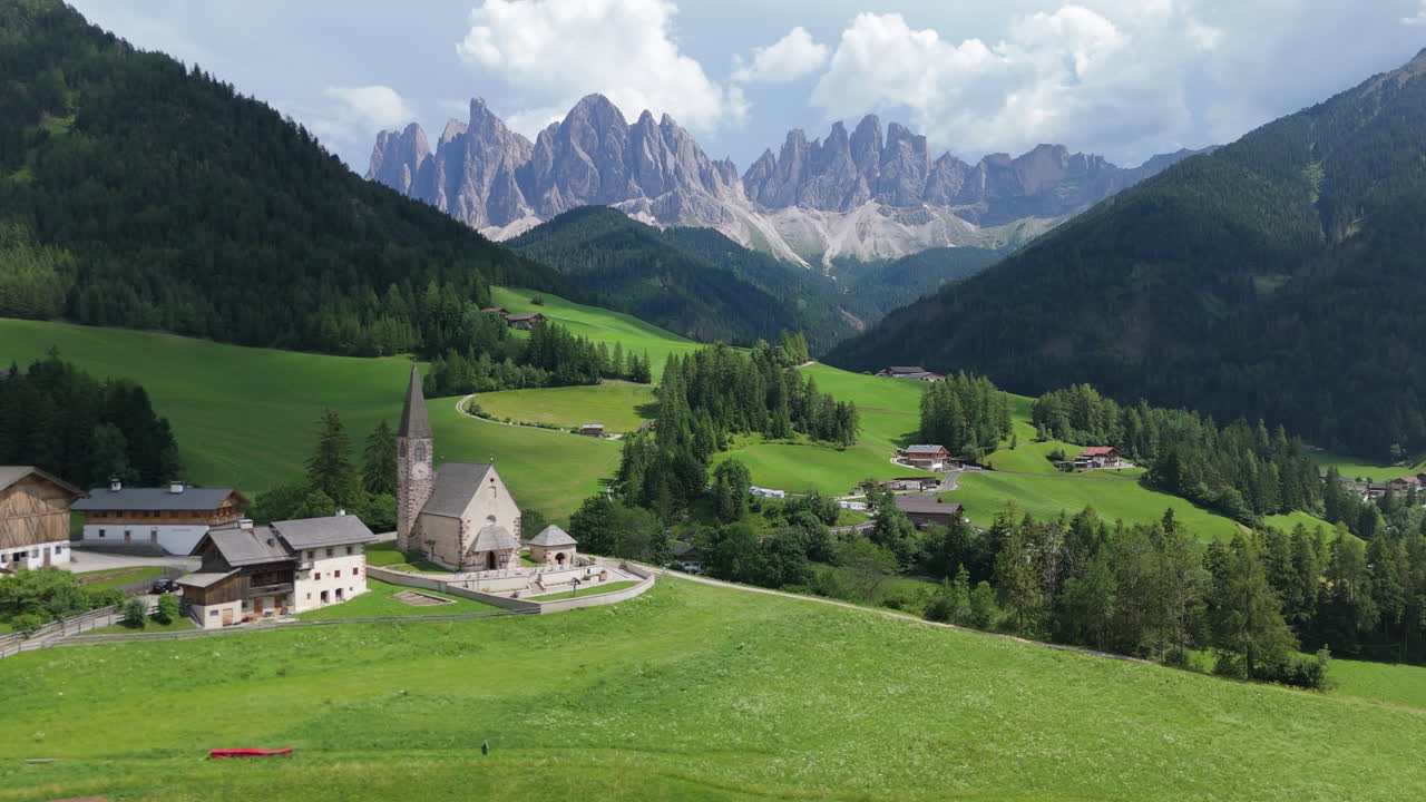Aerial view of the Dolomites with lush green alpine valley, small church and village houses framed by dramatic mountain peaks under summer sky