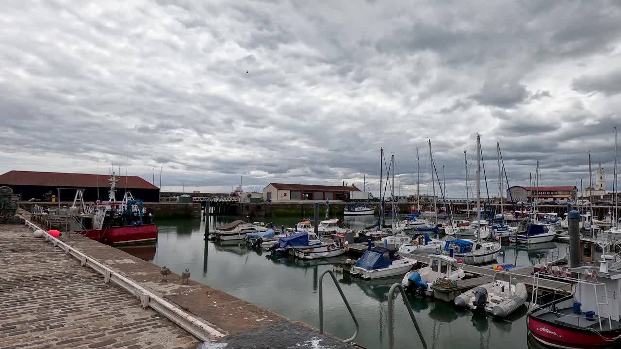 Camera moves along cobblestone dock toward moored boats under cloudy daylight at coastal marina