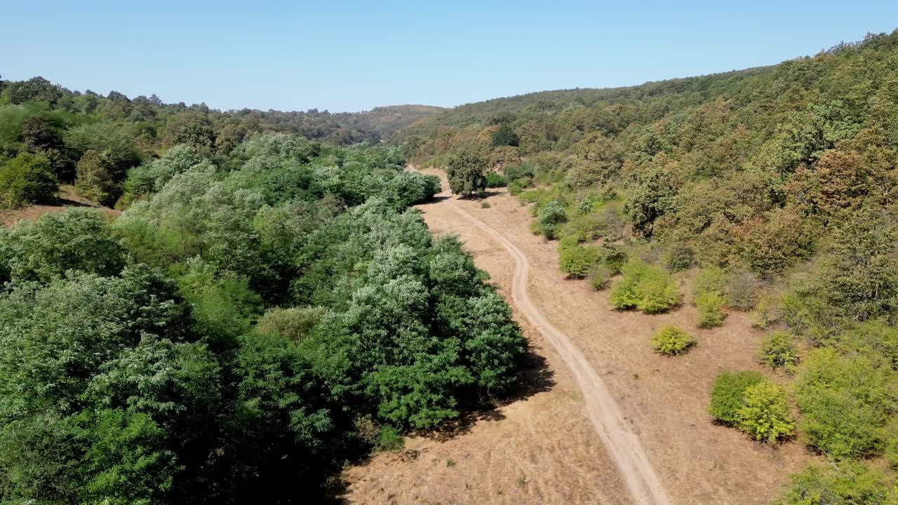 From a drone’s view, an old winding road cuts through a sweeping natural landscape, bordered by dense forests and open fields.