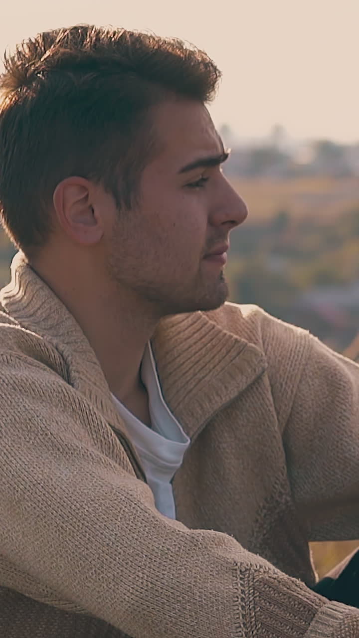 sad man in warm pullover sits suffering from headache with hangover on meadow lit by autumn sun closeup slow motion