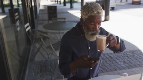 Afroamerikanischer-älterer-Mann,-Der-Sein-Smartphone-Benutzt,-Während-Er-Kaffee-Trinkt-Und-Draußen-Im-Café-Sitzt