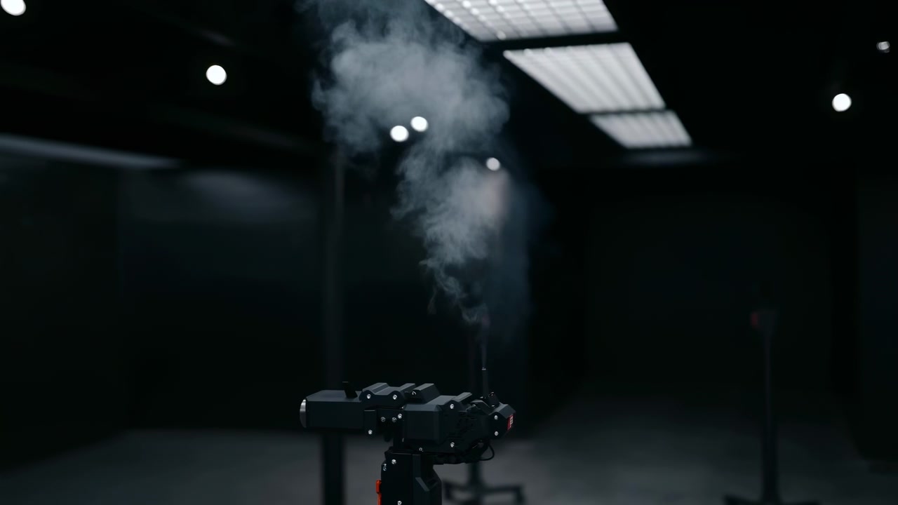 A black mechanical device sits in a dim industrial interior while vapor rises upward under overhead lights, creating a dramatic moody atmosphere