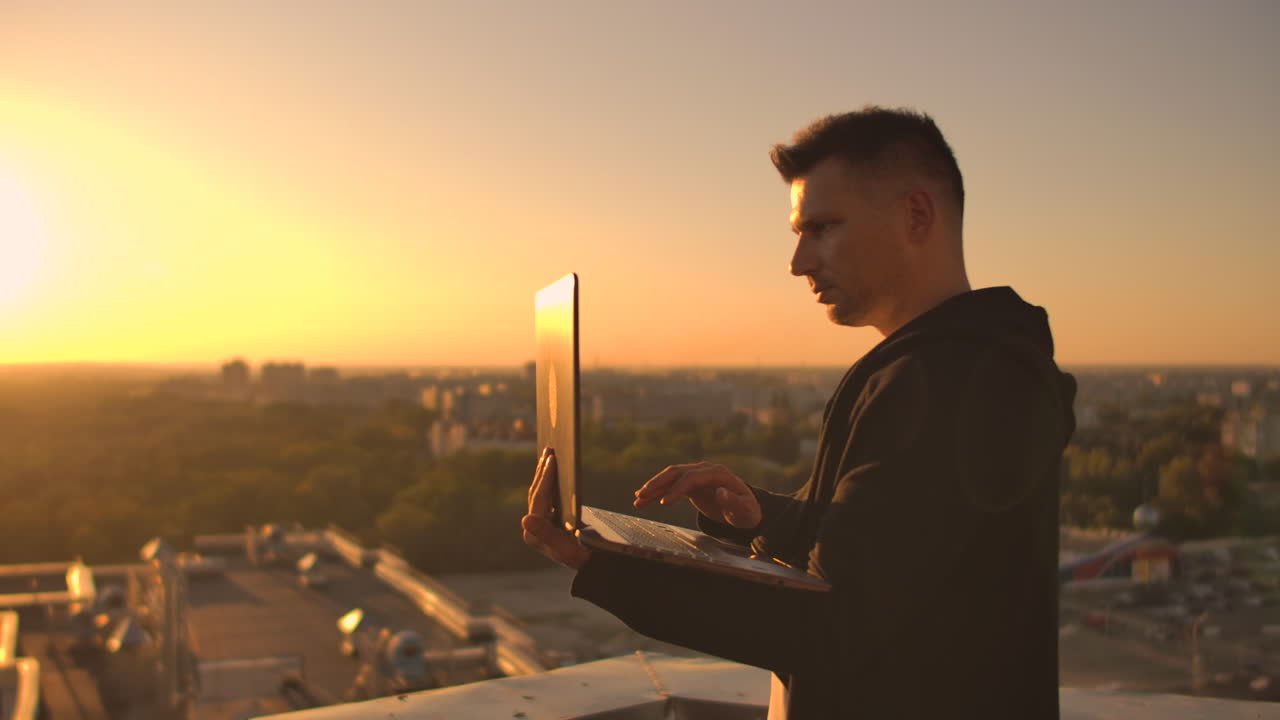 Middle plan: a male freelancer typing on a laptop keyboard at sunset overlooking the city while standing on the roof of a skyscraper. The businessman works remotely and controls the work of the company