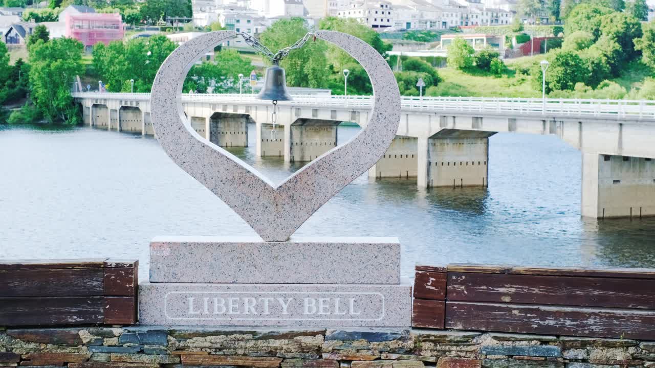 Static shot of the Liberty Bell in Portomarín on the Camino de Santiago. The bridge and river appear in the background. Symbolic composition blending history, journey and peaceful scenery