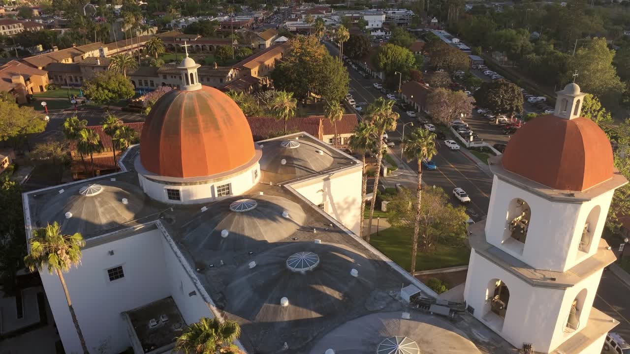 Ripresa aerea con drone in avanzamento della Missione Basilica San Juan Capistrano nella calda luce dell'ora d'oro, con la storica Missione di San Juan Capistrano visibile sullo sfondo, palme e il centro città.