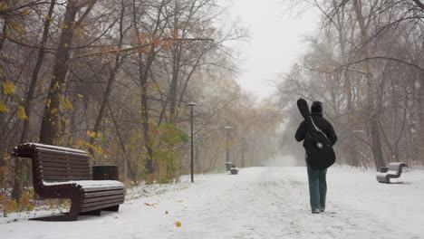 back view of a girl walking in a snowy park while carrying a guitar pack, winter trees and benches lining the pathway, capturing a serene snowy day