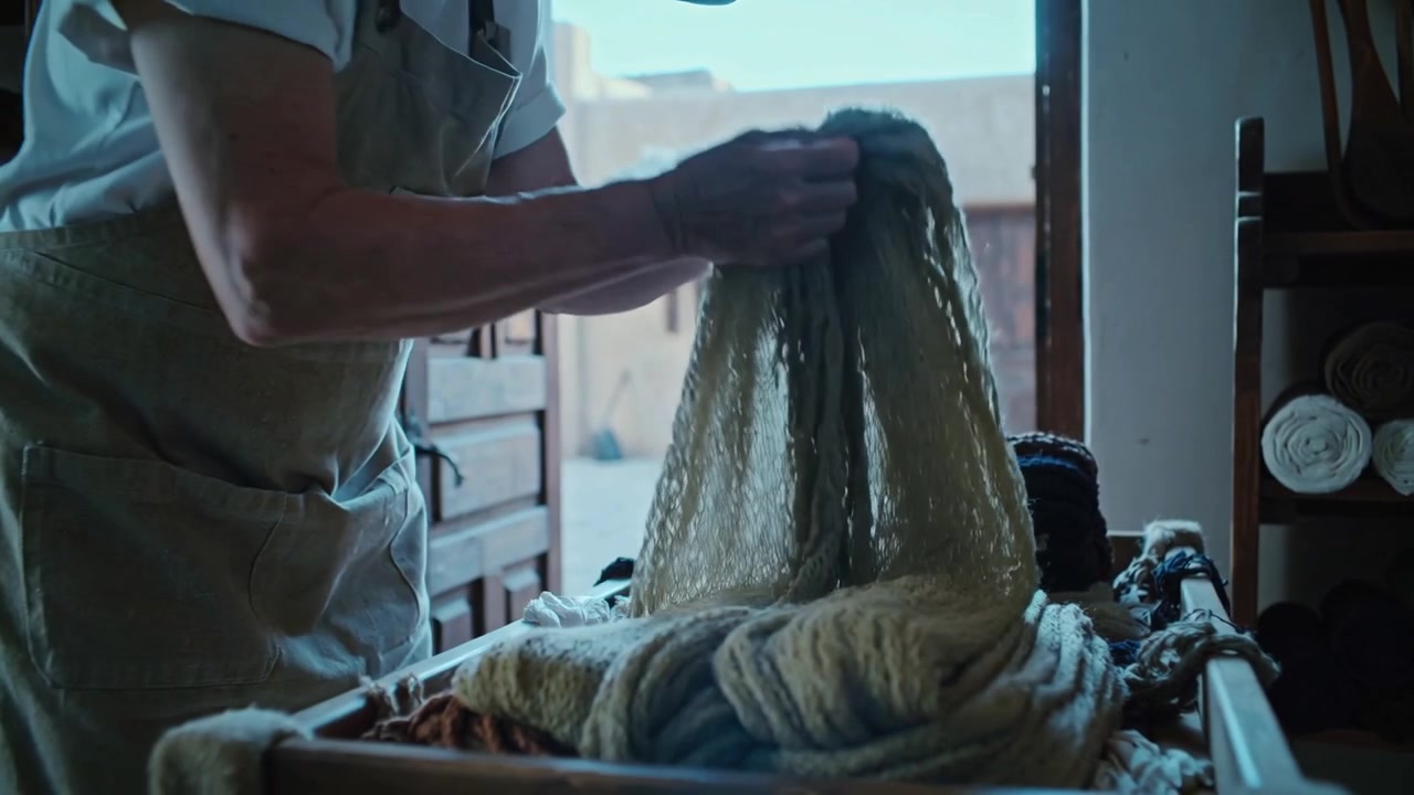 Standing at low cart, vendor wearing apron and hat smoothing woolens in shop to organize display