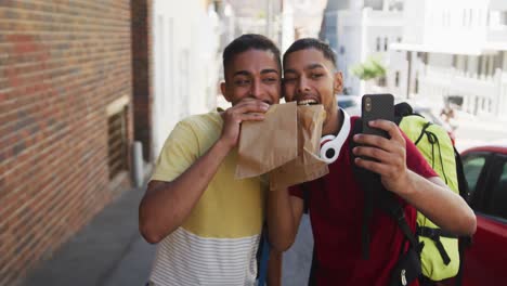 Dos-Amigos-Felices-De-Raza-Mixta-Comiendo-Y-Tomándose-Selfie-En-La-Calle