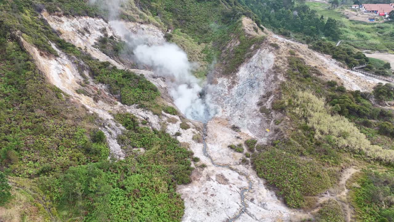 Drone video of hot steam rising from a sulfur-rich crater, surrounded by rocky hills and patches of green vegetation in a volcanic region