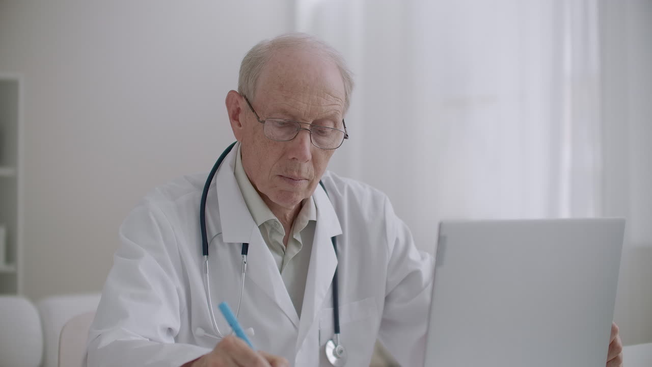 un anziano professore di medicina sta guardando un webinar tramite laptop nell'ufficio della clinica e sta prendendo appunti e-learning per i professionisti della salute