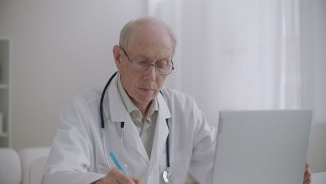 un profesor de medicina de edad avanzada está viendo un seminario web por ordenador portátil en la oficina de la clínica y tomando notas de aprendizaje electrónico para profesionales de la salud