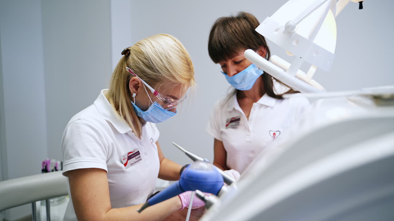 Female dentist wearing mask and protective goggles providing dental care for the patient. Assisting nurse stands beside.