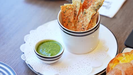 close-up of crispy herb crackers served with a green pesto dip on a doily-lined plate. ideal for food, fine dining, or menu content