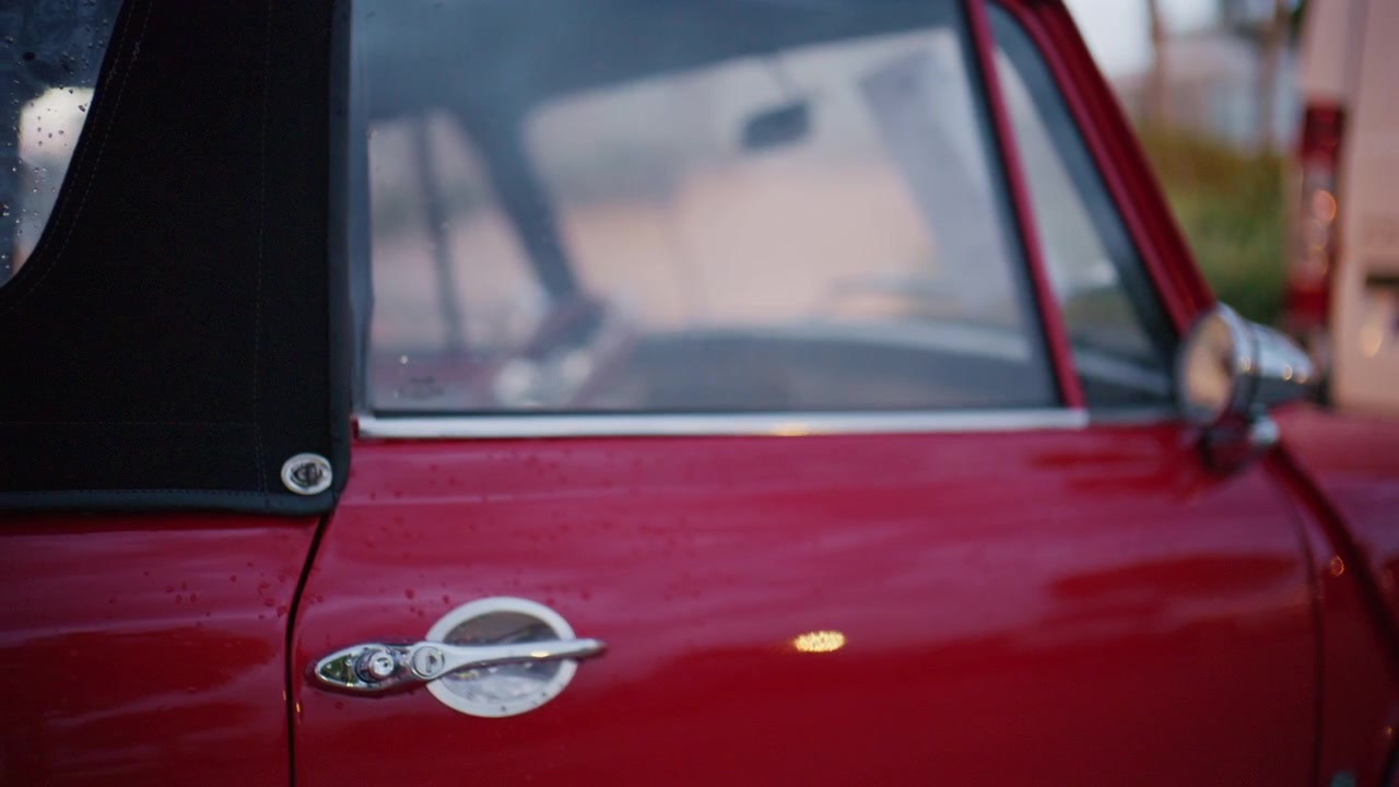 Primo piano della portiera di un'auto decappottabile rossa d'epoca con maniglia cromata, capote nera e gocce di pioggia