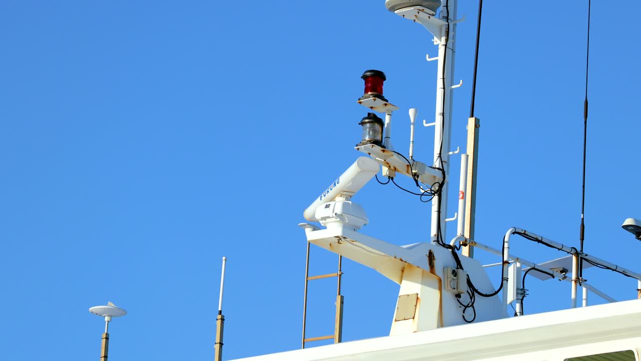 Radar and antennas on a ship's mast rotate against a clear blue sky. Bright daylight illuminates the scene, highlighting maritime technology