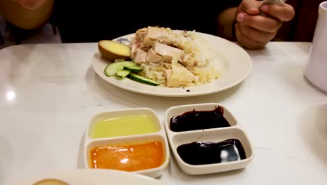 A person uses chopsticks to pick up steamed chicken from a rice plate, dipping it into three different sauces on a bright, clean tabletop