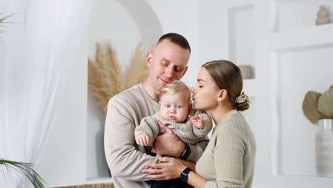 portrait of a happy caucasian family standing together in the room. loving parents lean and kiss their baby.