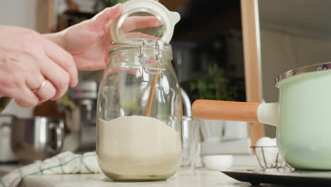 hand view of person opening glass container filled with flour, using wooden spoon to scoop and shake contents for accurate measurement, with visible flour dust rising in soft natural kitchen