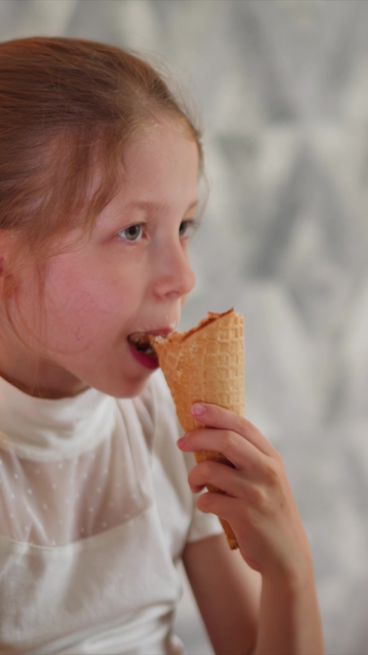 Close up of young girl eating ice cream cone while holding it in hand. Girl wears white blouse with polka dots against soft gray patterned background. Ice cream is partially eaten waffle