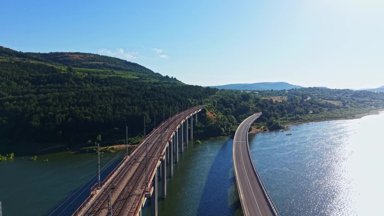 Un lungo ponte attraversa un fiume, affiancato da colline alberate, sotto la luce brillante del sole durante il giorno.