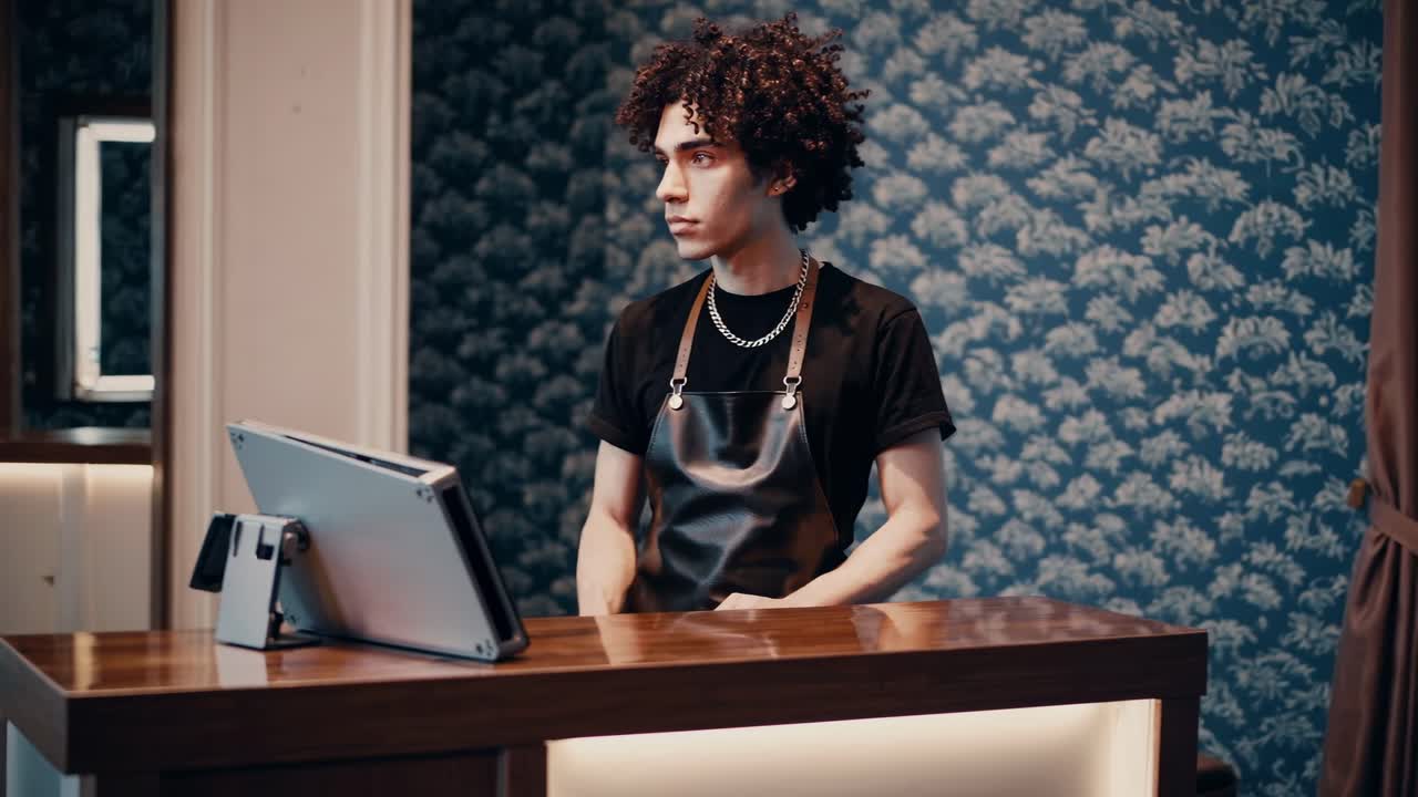 Young professional hairdresser wearing black apron standing at reception desk in modern beauty salon, working on tablet while waiting for incoming clients during workday