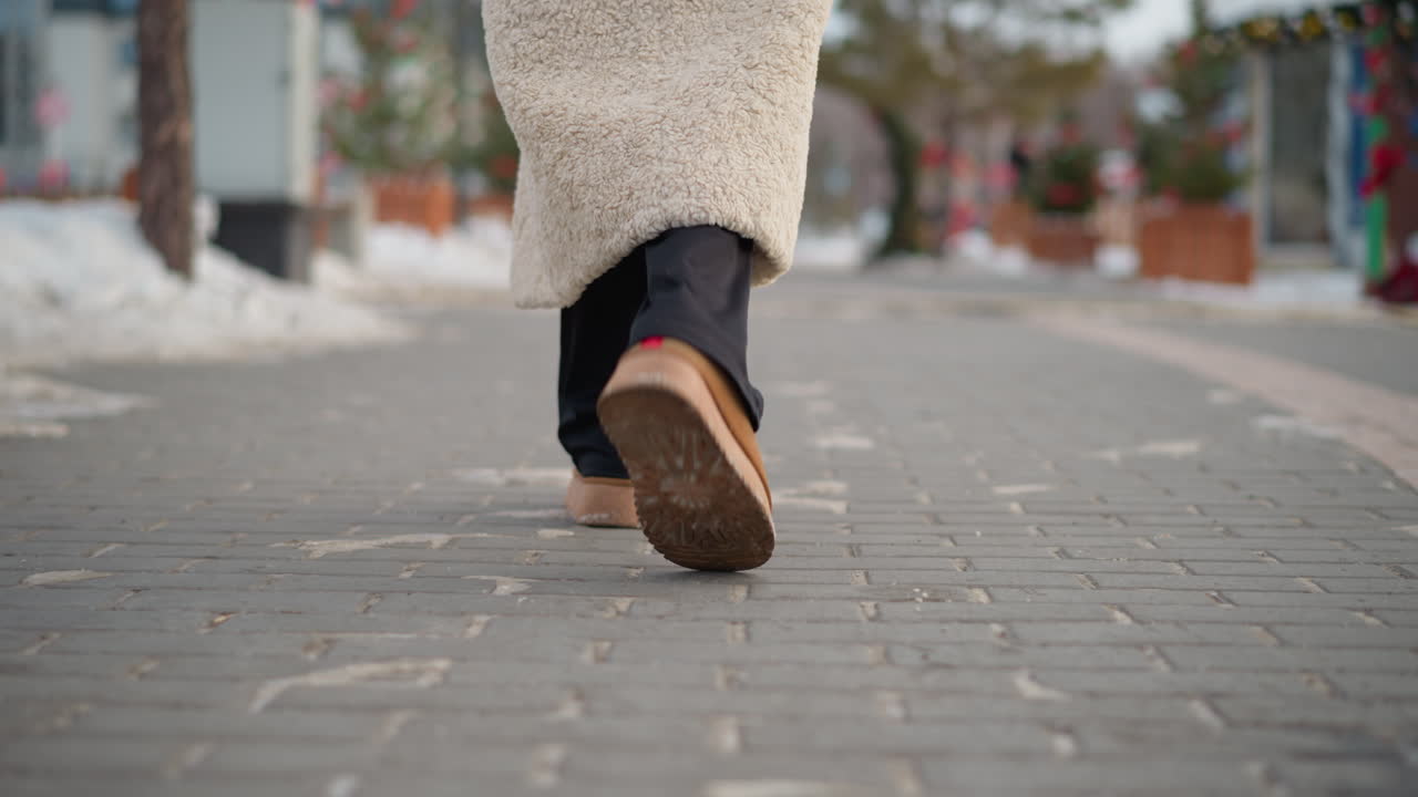 vista de fondo de cerca de una dama con un cálido abrigo de invierno beige y botas de gamuza marrón caminando por una calle pavimentada y nevada, el fondo borroso con flores y árboles decorados agrega una atmósfera festiva y acogedora