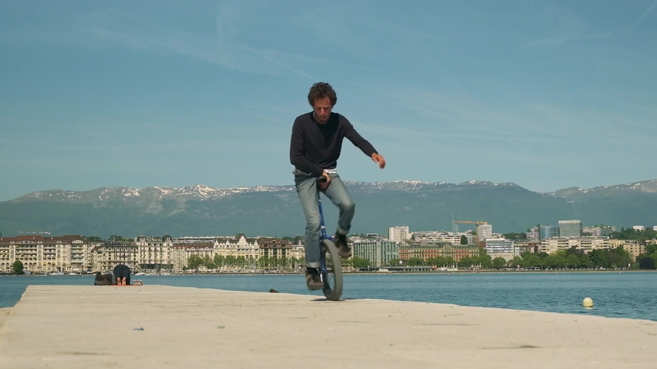 Uomo esegue acrobazie in monociclo su una passeggiata lungomare con vista sulla città e sulle montagne.