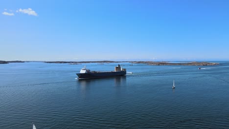 Buque-De-Carga-Navegando-En-El-Océano-Con-Veleros-Navegando-Tranquilamente-Durante-El-Verano-En-Lysekil,-Suecia