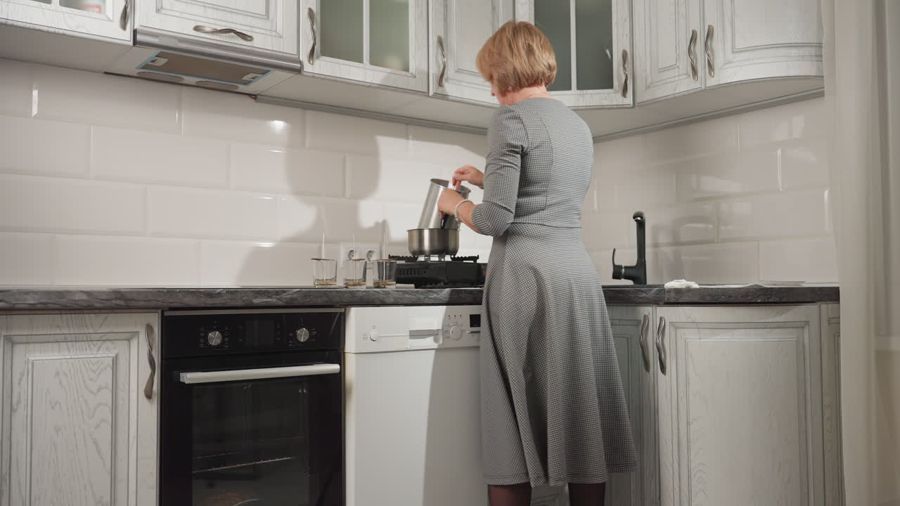 Back view of woman in patterned dress cooking at kitchen counter with three empty glass cups arranged neatly beside stove, showing calm focused environment