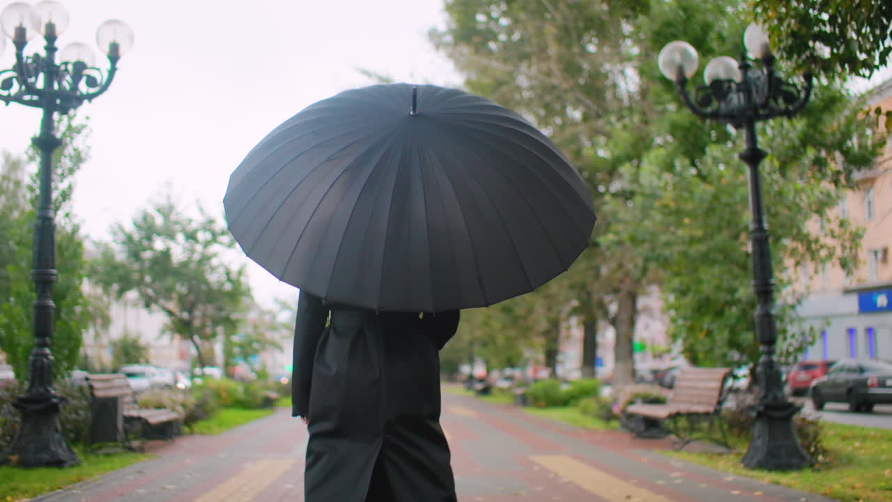 Person dressed in black holding large umbrella walking along urban park pathway on cloudy autumn day with benches, trees, street, and cars in background creating moody calm atmosphere of solitude