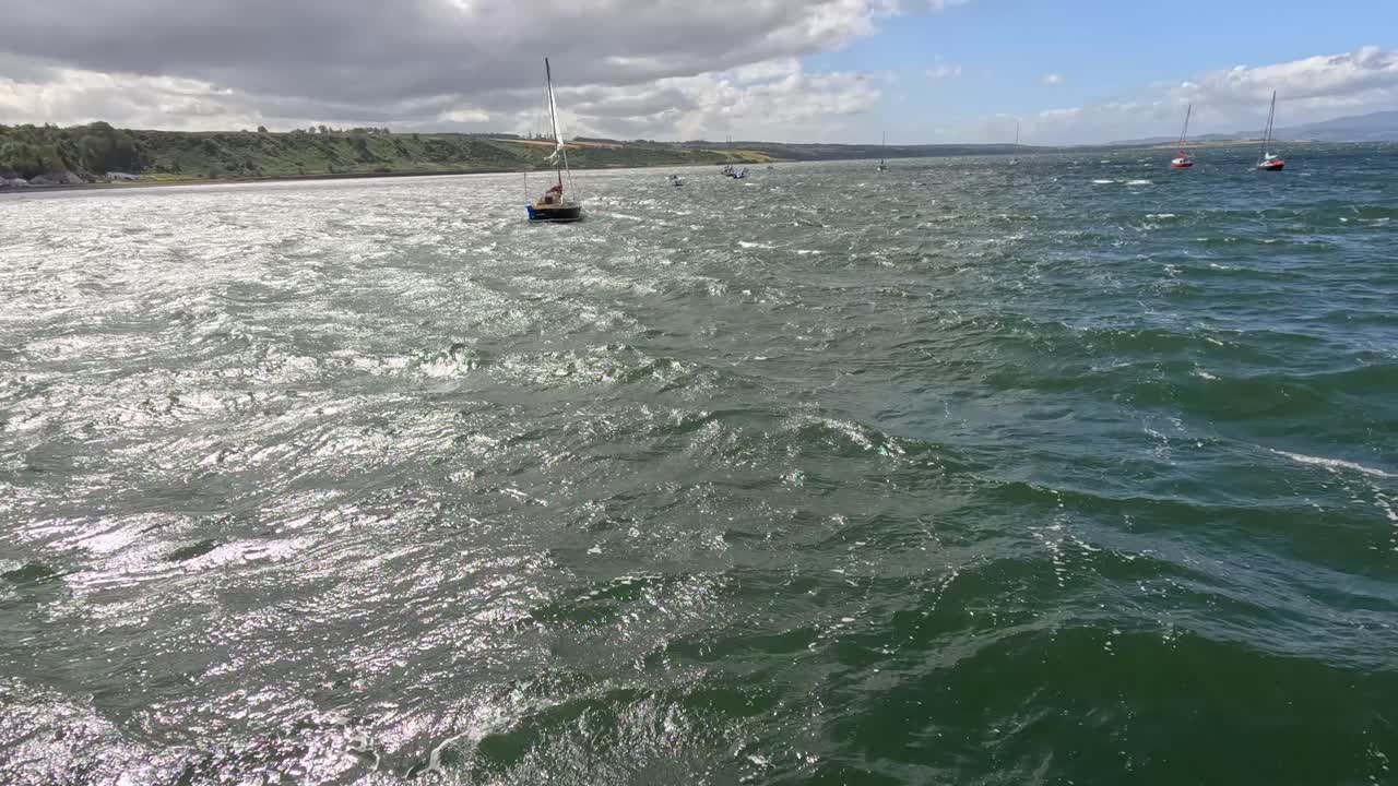 A single sailboat floats on choppy, sunlit waters near the Cromarty coastline. The camera glides steadily over the sea, capturing dynamic reflections and distant shoreline