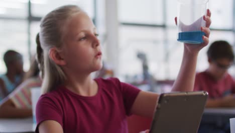 colegiala caucásica usando una tableta mirando líquido azul en un frasco durante la clase de química