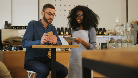 Joven-Caucásico-Haciendo-Un-Pedido-En-El-Café,-Mientras-Que-La-Testigo-Afroamericana-Escribe-Todo-En-Una-Tableta